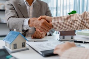 Two people in business attire finalize a real estate deal, shaking hands across a desk with house models, keys, and a contract, symbolizing agreement, trust, and successful negotiation.